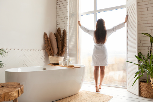 Morning Beauty Routine. Woman Opening Windows In Bathroom Standing Back To Camera Wearing White Bathrobe Preparing For Domestic Spa Rituals In Modern Bathing Room At Home.